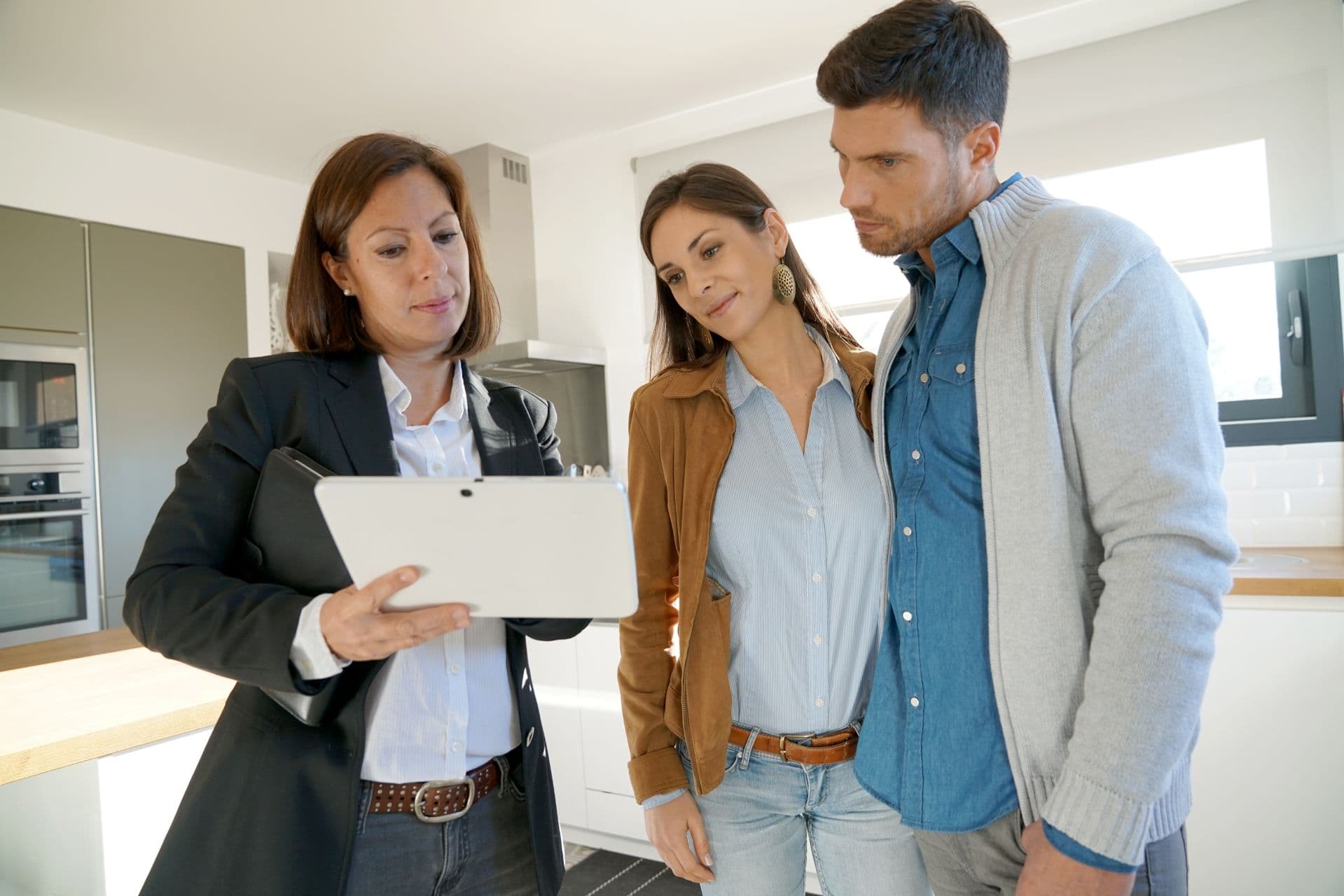 A real estate agent in a blazer shows a couple a tablet screen in a modern kitchen. They look engaged and thoughtful, discussing details.