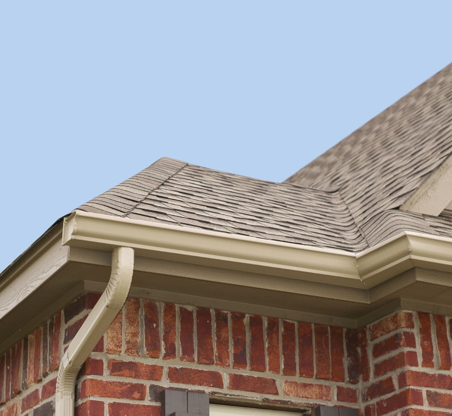 Close-up of a house corner with beige roof shingles, a brick wall, and beige gutters under a clear blue sky.