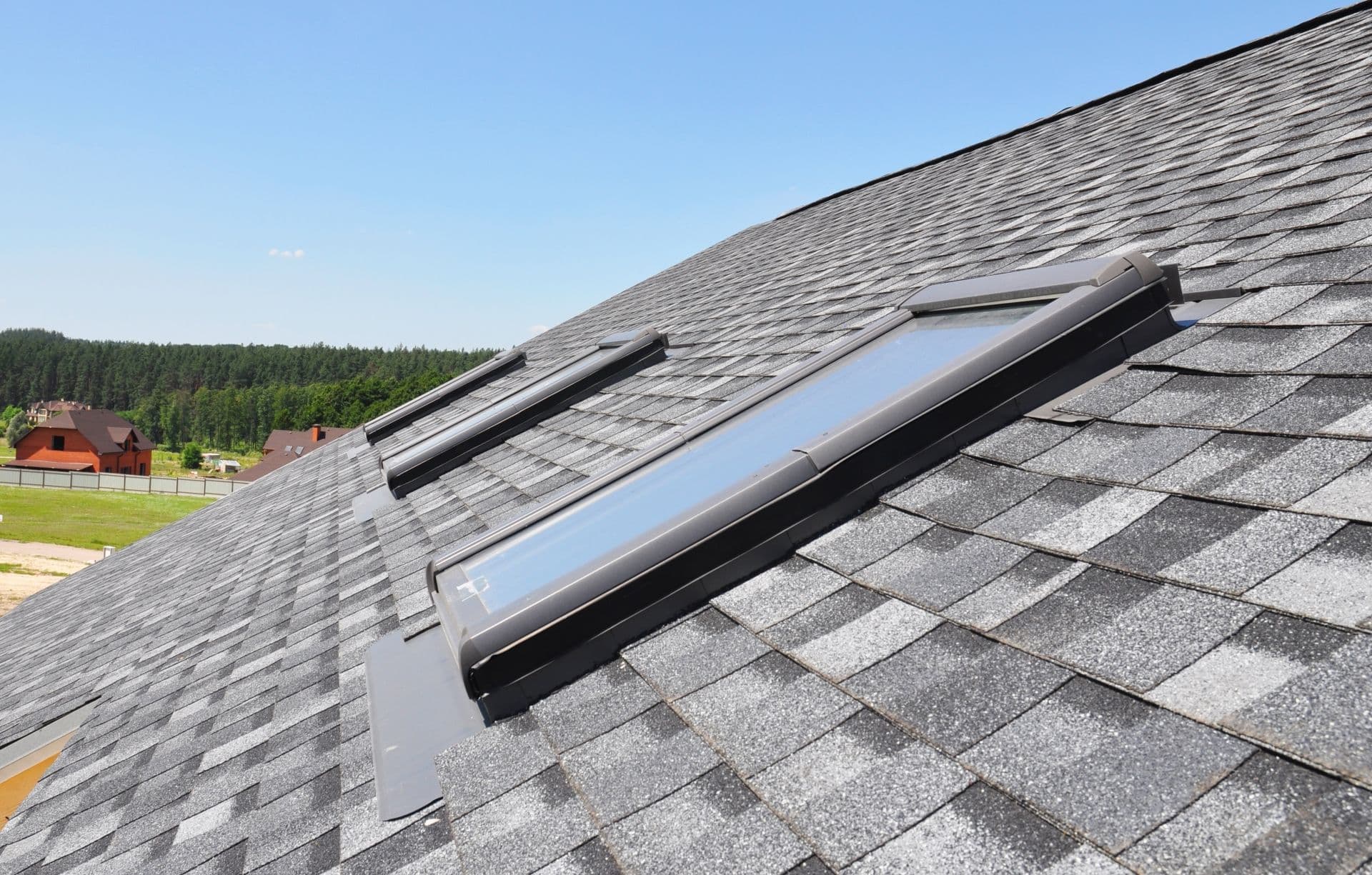 Gray shingle roof with three skylights under a clear blue sky. In the background, there are green trees and a red house.