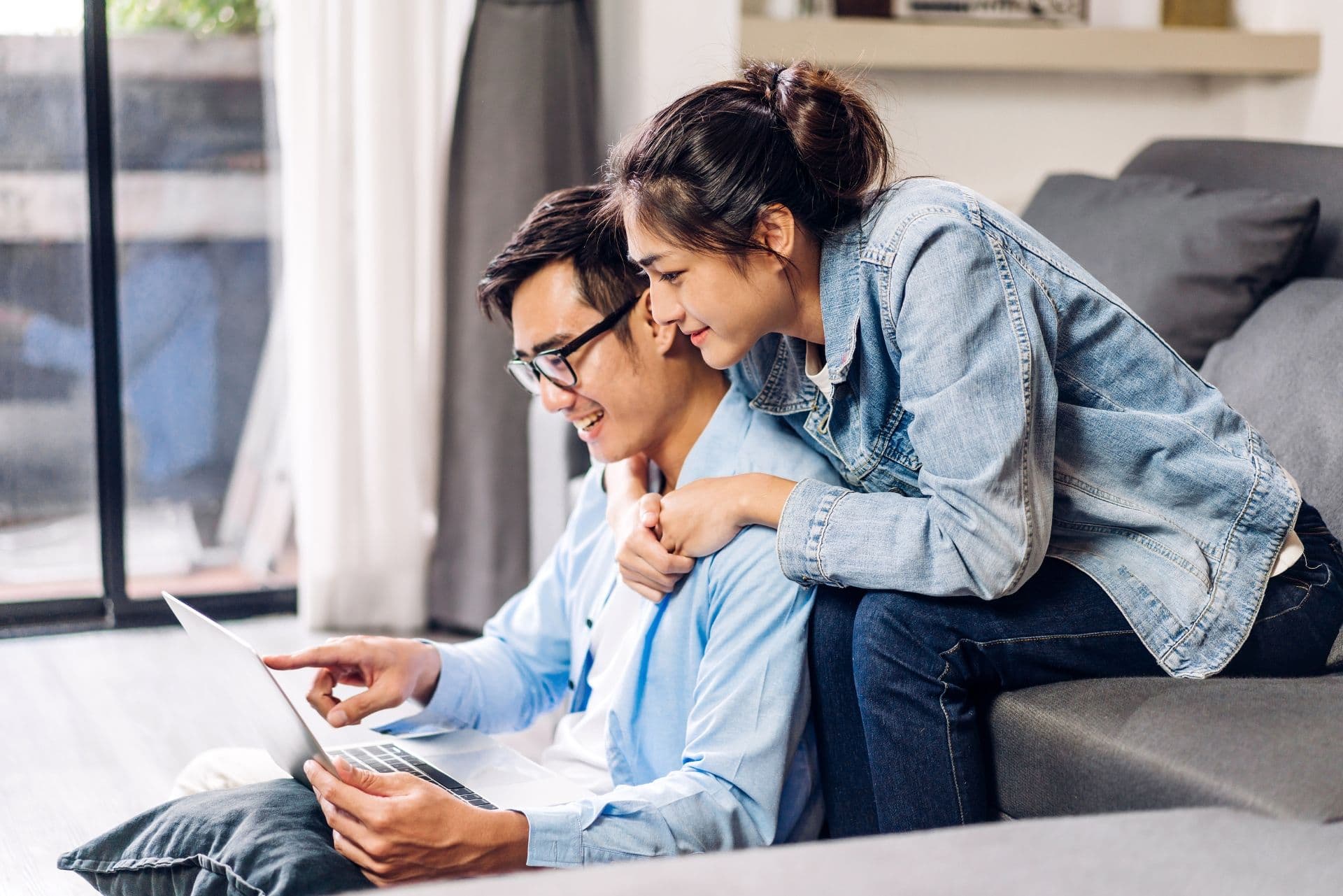 A couple sits on the floor, smiling and looking at a laptop. The man points at the screen while the woman, in a denim jacket, leans over him.
