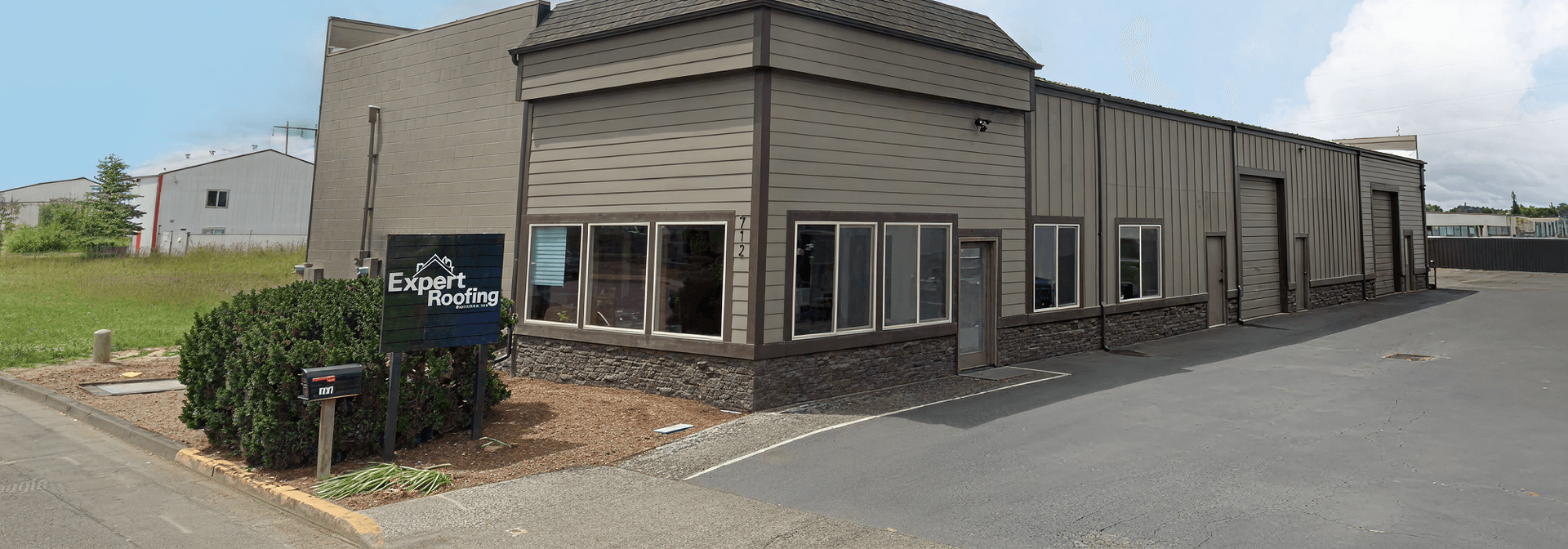 Front view of a modern commercial building with gray siding and stone accents. A sign reads "Expert Roofing."