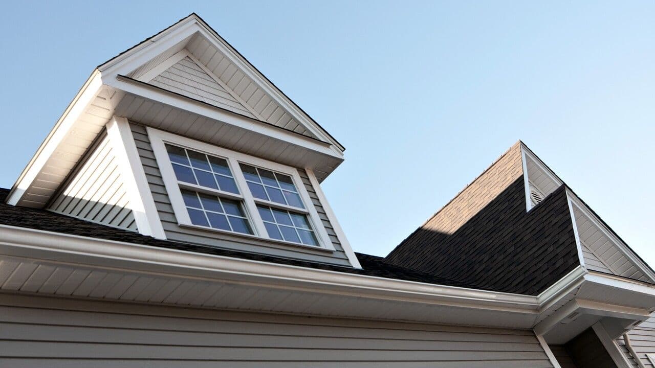 A house roof featuring a dormer with gray siding, double-pane windows, and white trim under a clear blue sky.
