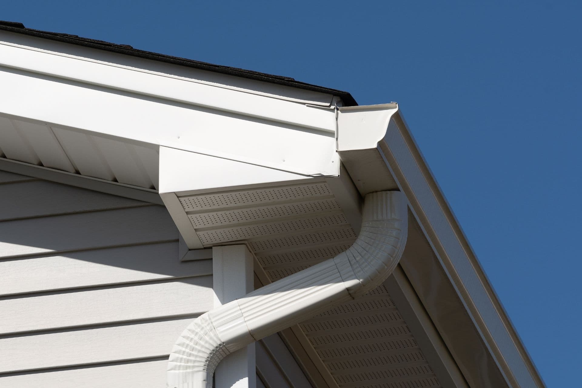 Corner of a house with beige siding and a white gutter system against a clear blue sky. The gutter curves downward.
