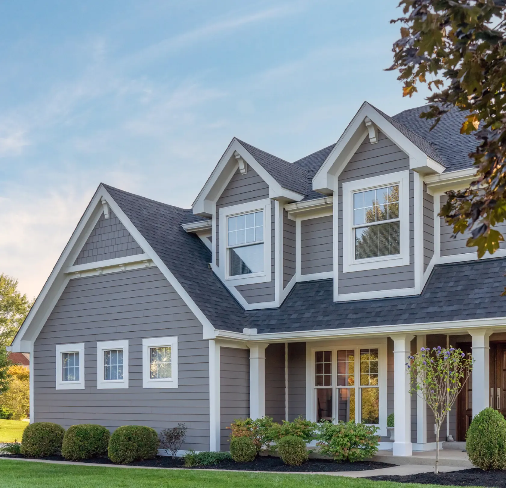 Two-story gray house with white trim and multiple peaked roofs. Neatly trimmed shrubs line the front.