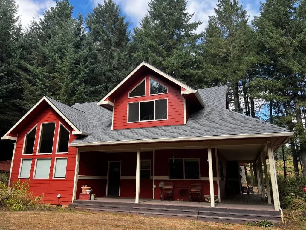 A charming, red two-story house with large windows and a new gray shingle roof is nestled amongst tall pine trees.