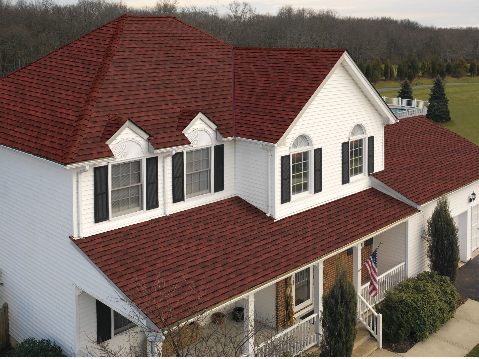 A white two-story house with a vibrant red shingle roof stands in a scenic area. Black shutters adorn the windows, and a U.S. flag hangs out front.