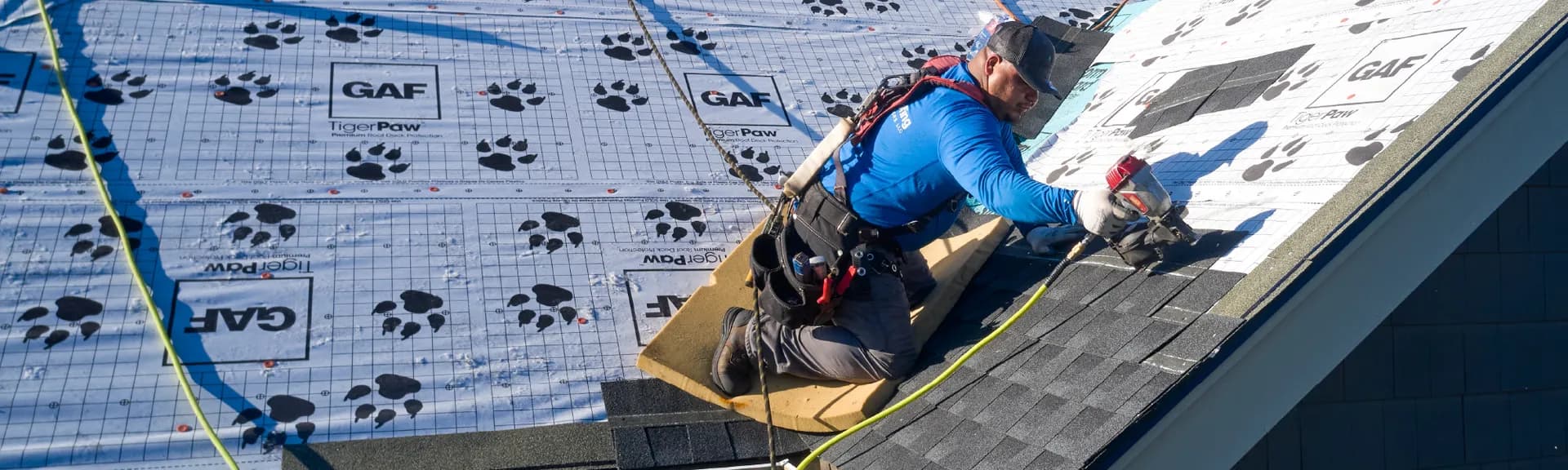 A worker in a blue shirt and harness kneels on a sloped roof, placing asphalt shingles over a pattern-marked underlayment.