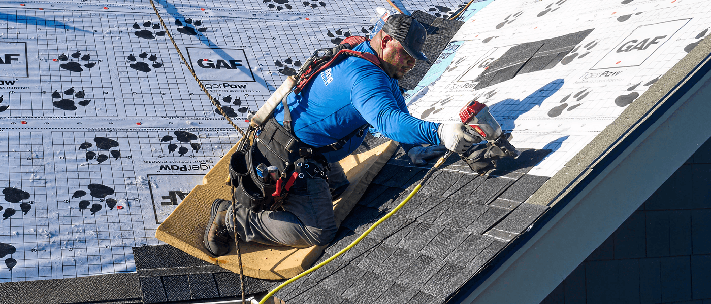 A worker in a blue shirt and harness kneels on a sloped roof, placing asphalt shingles over a pattern-marked underlayment.