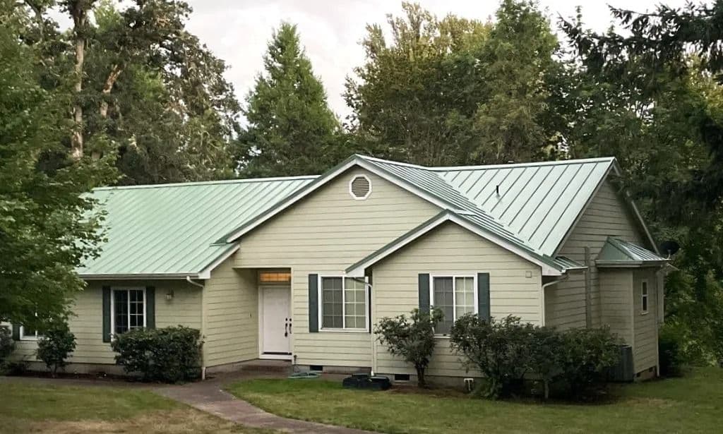 Single-story beige house with green metal roof, surrounded by lush trees and shrubs.