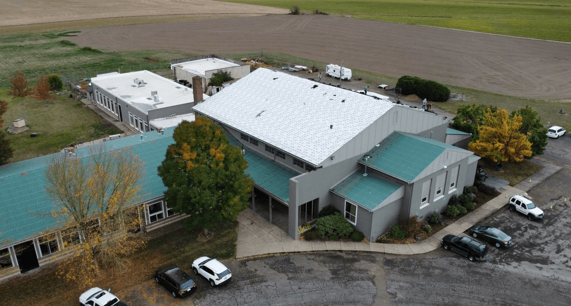 Aerial view of a large, L-shaped building with a white roof and green awnings, surrounded by trees with autumn foliage. Vehicles are parked nearby.