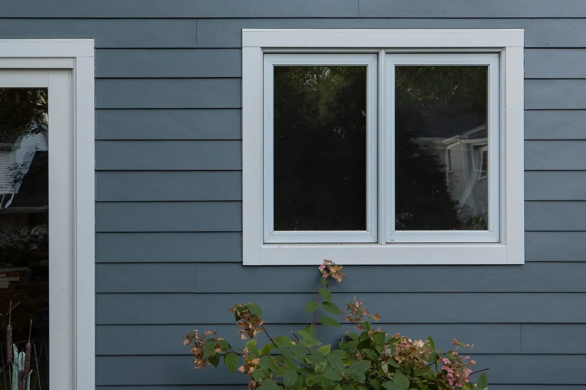 Blue house exterior with horizontal siding, featuring a white-framed window and door. In the foreground, a flowering bush adds a natural touch.
