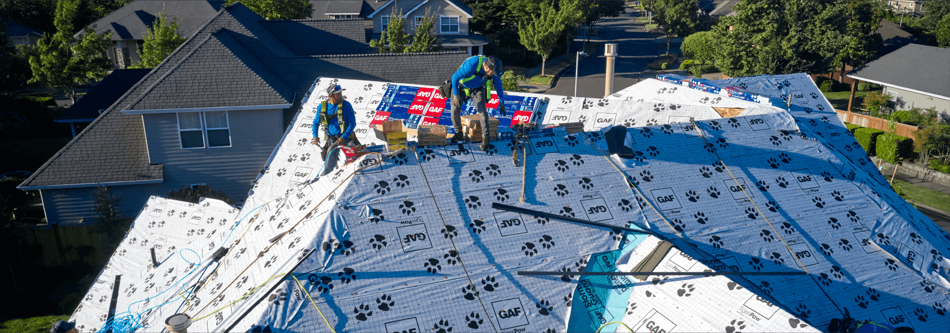 Workers wearing safety gear install roofing underlayment on a residential roof in a suburban neighborhood, with trees and houses in the background.