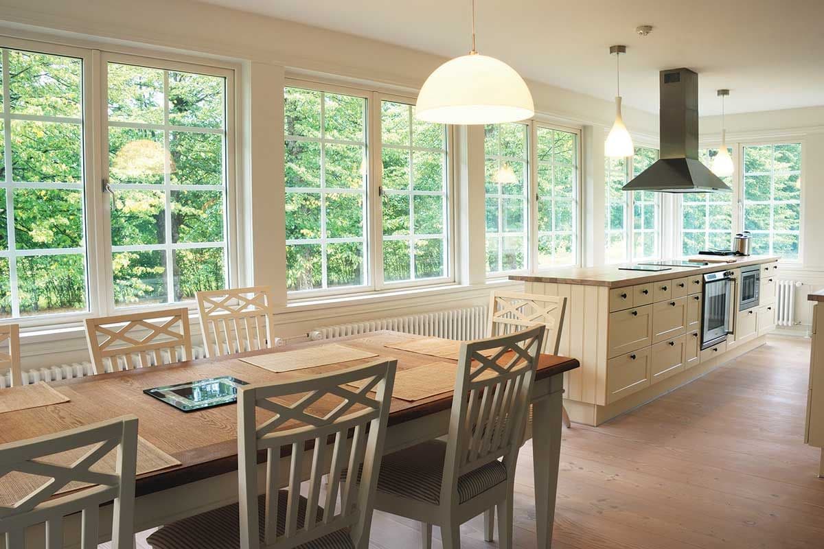 Bright kitchen with large windows overlooking lush greenery. Features a wooden dining table, chairs, and a modern island with pendant lights.