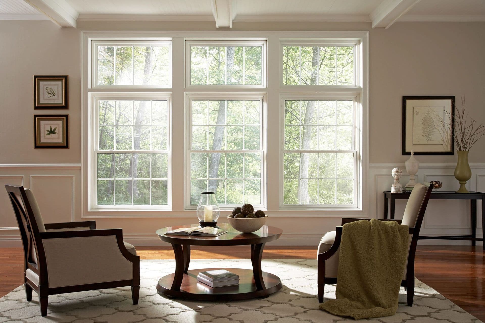 Spacious, sunlit living room with large windows showcasing lush greenery. Features two armchairs, a round coffee table, and elegant decor.