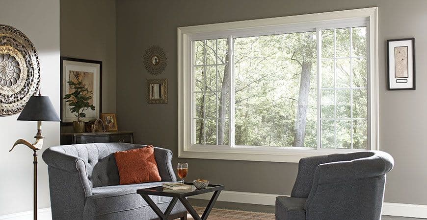 Cozy living room with gray chairs and a small table, featuring a large window showcasing lush green trees.