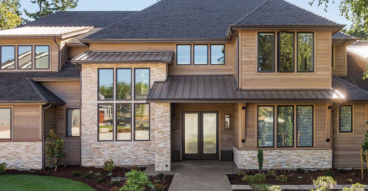 Modern two-story house with a gabled roof, large windows, stone and wood exterior. A welcoming front yard with small plants and a concrete pathway.