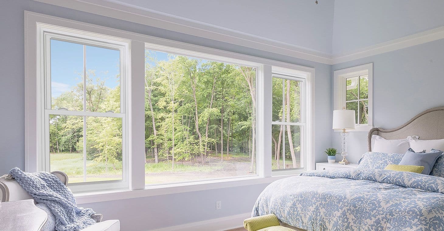 Bright bedroom with large windows showcasing a lush green forest. The space features light blue walls, a cozy bed with floral bedding, and a serene ambiance.